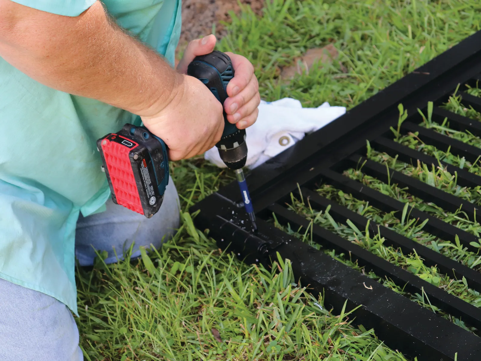 A person kneels on grass, using a power drill to assemble a black metal fence panel.