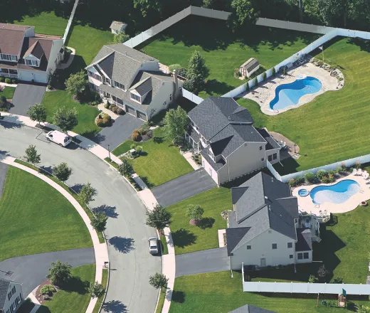 Aerial view of suburban neighborhood featuring curved street, multiple houses with well-kept lawns, and two properties with kidney-shaped swimming pools in fenced backyards. Surrounding greenery and trees are visible.